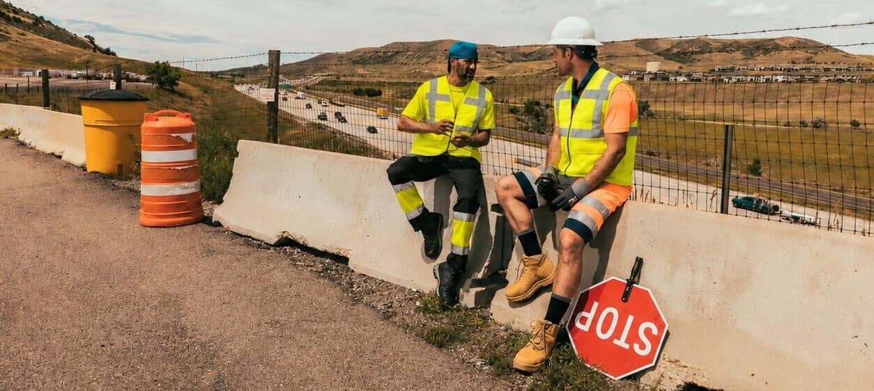 Due operai sono seduti su una barriera di cemento a lato della strada. Indossano indumenti ad alta visibilità a maniche corte e gilet rinfrescanti. Sullo sfondo, si può vedere un paesaggio montano brullo e un cielo azzurro nuvoloso.