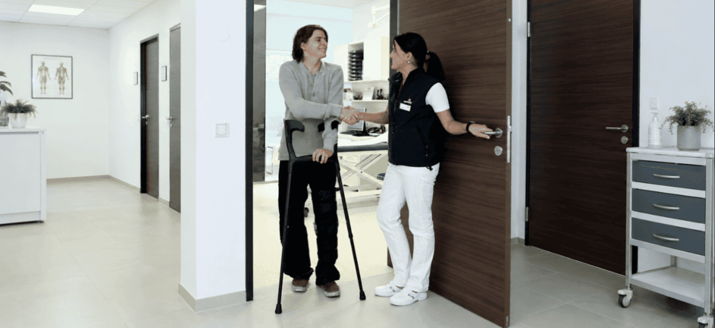 Due persone sono ritratte nel corridoio di una clinica. Un uomo con le stampelle stringe la mano a una dottoressa.