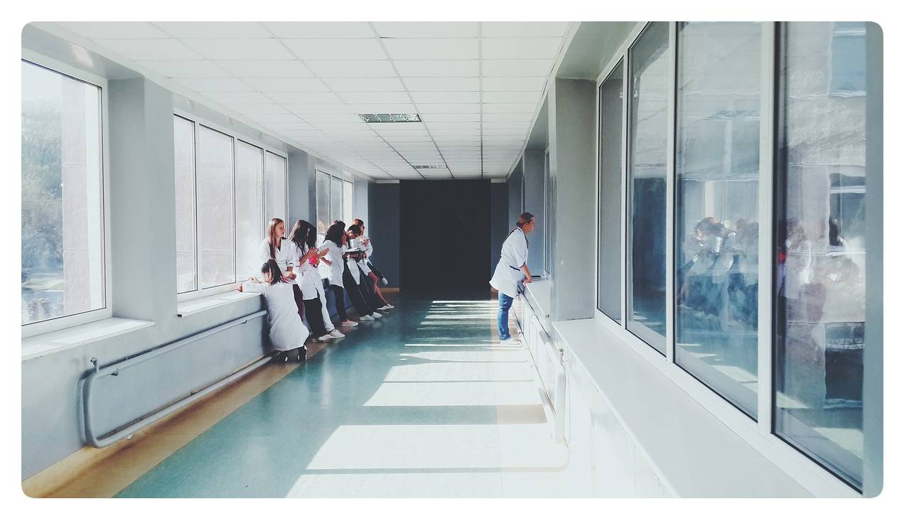 Si vede un corridoio con finestre a destra e a sinistra, in cui un gruppo di operatori sanitari aspetta appoggiato alla parete.