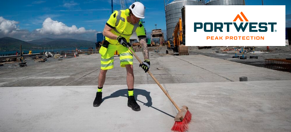 Un uomo con tuta da lavoro gialla ad alta visibilità e casco bianco sta spazzando il pavimento di cemento davanti a una fabbrica. Sullo sfondo si vedono montagne e un cielo azzurro con piccole nuvole. Il logo blu e arancione del produttore, Portwest, è posizionato nell'angolo in alto a destra.