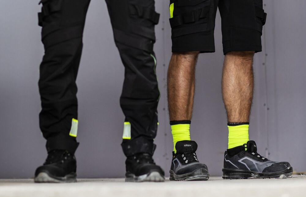 Si vedono due paia di gambe maschili: un uomo indossa un pantalone da lavoro lungo nero, l&rsquo;altro un pantaloncino da lavoro corto giallo ad alta visibilit&agrave;. &Egrave; presente un link alla nostra categoria di pantaloni da lavoro.