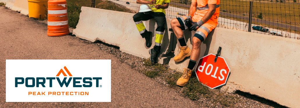 Si possono vedere le gambe di due persone in abiti da lavoro. Sei seduto su una barriera di cemento, c'è un segnale di stop accanto a te e sullo sfondo ci sono una recinzione, una strada e una catena di colline. Nell'angolo in basso a sinistra dell'immagine si trova il logo del produttore Portwest in arancione e blu su sfondo bianco.
