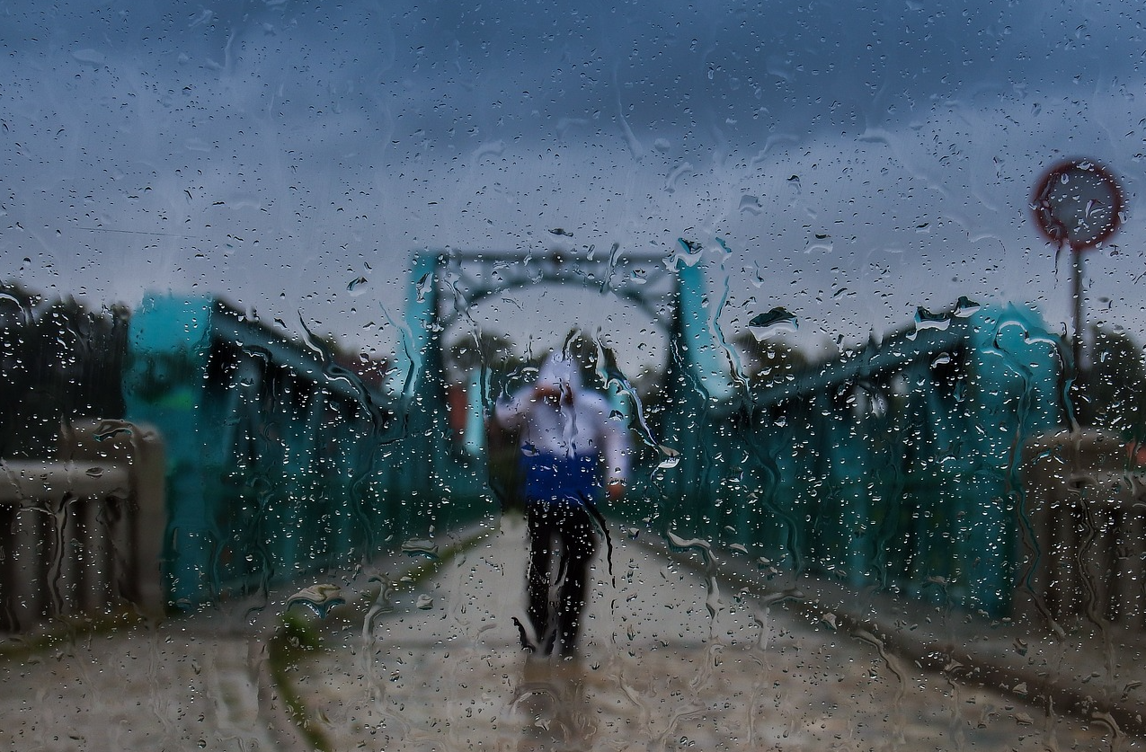 Ponte blu contro un cielo grigio con una persona in tenuta da pioggia in mezzo alla strada. La scena è stata fotografata attraverso una lastra di vetro bagnata. Viene fornito un collegamento alla nostra intera selezione di pratici indumenti antipioggia.