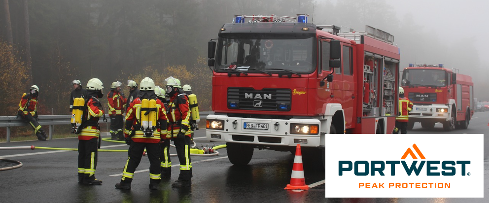 Scena di strada con due autopompe rosse e vari vigili del fuoco. Sullo sfondo si vedono alberi coperti di nebbia. Nell'angolo in basso a destra c'è il logo Portwest blu-arancione davanti a un rettangolo bianco.