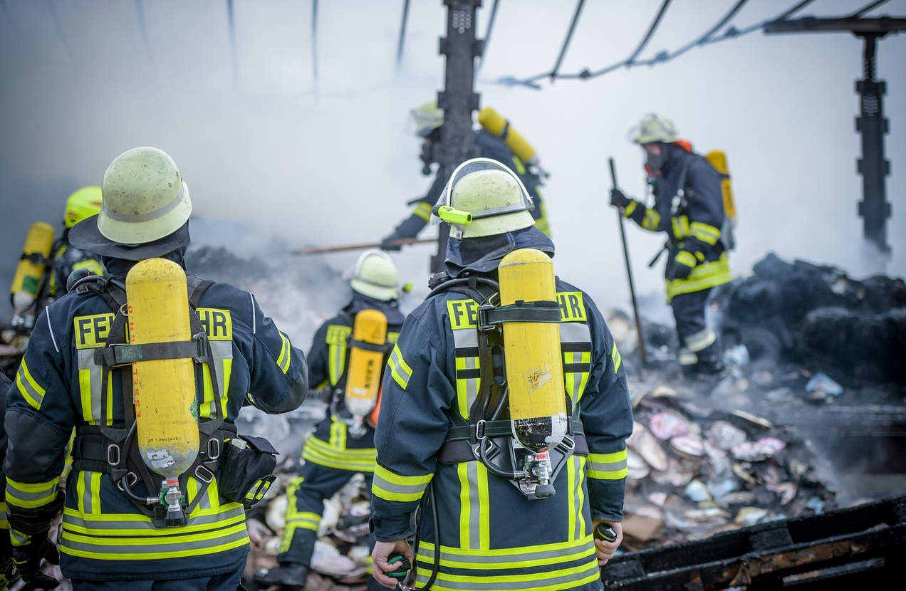 Vigili del fuoco in abiti blu con strisce di avvertimento gialle e caschi in un deposito di rottami. Il collegamento fornito conduce alla nostra selezione di indumenti antincendio ignifughi.