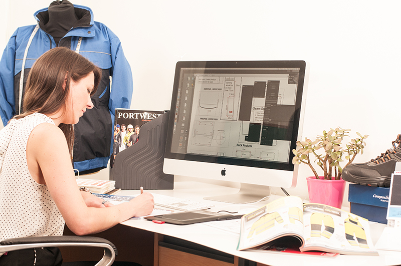 Bruna donna che lavora alla scrivania. Sullo schermo del suo computer si possono trovare disegni tecnici di abiti da lavoro.