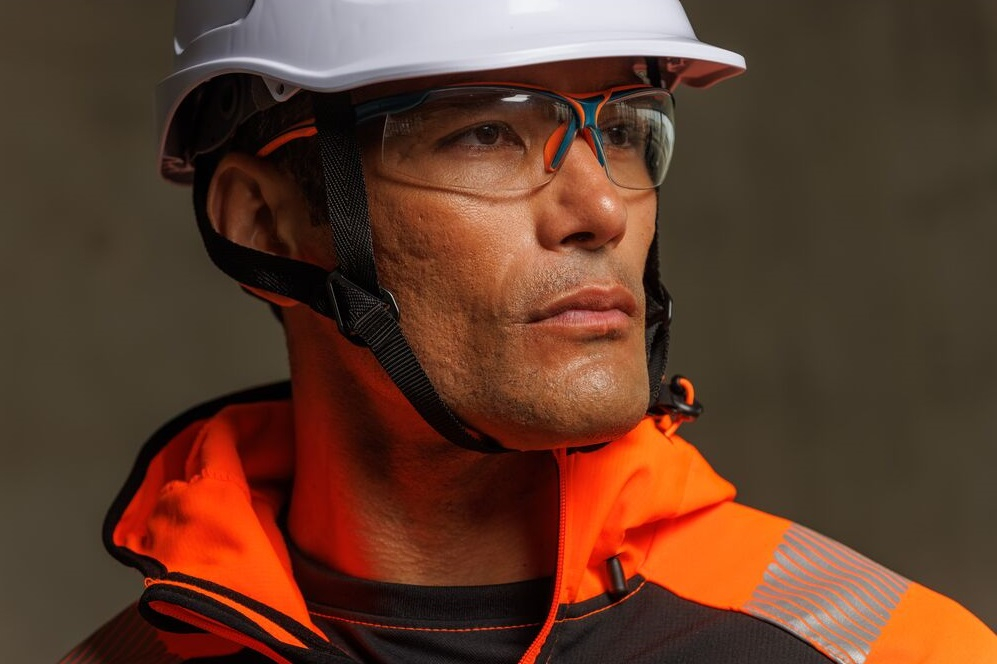 Primo piano di un operaio con casco protettivo bianco e occhiali di sicurezza trasparenti, che guarda concentrato in lontananza; indossa abbigliamento ad alta visibilità arancione e nero. Collegamento alla nostra collezione di protezione per gli occhi incluso.