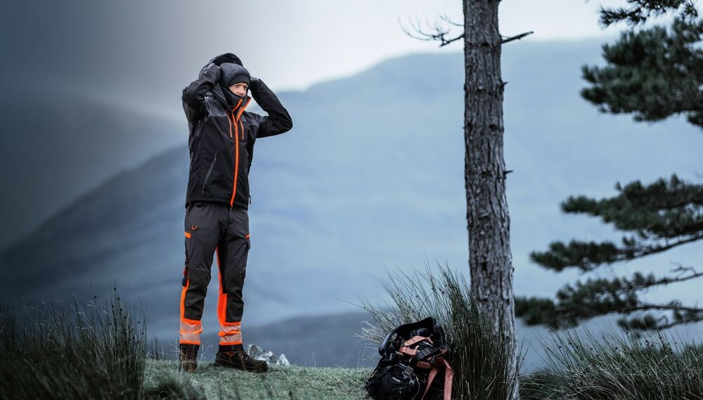 Lavoratore con abbigliamento da lavoro impermeabile con elementi riflettenti è all'aperto in montagna e si mette un berretto invernale. Un link ai nostri berretti è incluso.