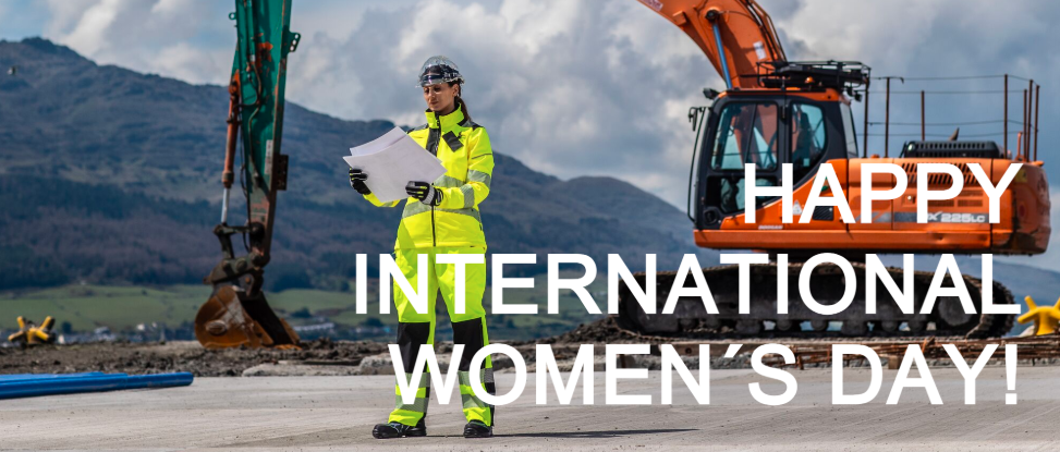 L'immagine mostra una donna in un cantiere edile con indosso un equipaggiamento di sicurezza giallo brillante, tra cui casco e occhiali protettivi, e con in mano delle planimetrie. Dietro di lei c'è un grande escavatore arancione con il braccio alzato e sullo sfondo si estendono verdi colline sotto un cielo parzialmente nuvoloso. Sopra l'immagine c'è scritto a grandi lettere bianche "BUONA GIORNATA INTERNAZIONALE DELLA DONNA!" in inglese.