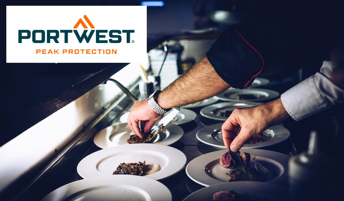 Due chef, ciascuno con un braccio in giacca da cuoco nera e l'altro in giacca da cuoco bianca, dispongono con cura il cibo su piatti bianchi in una cucina professionale. In alto a sinistra è visualizzato il logo Portwest "Peak Protection". C'è un collegamento che porta agli abiti del nostro chef.