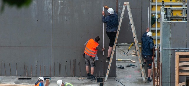 Due operai sono raffigurati in un cantiere. Uno è in piedi su una scala, l'altro la tiene ferma. Entrambi indossano pantaloncini da lavoro; uno indossa un casco bianco e l'altro un gilet arancione ad alta visibilità.