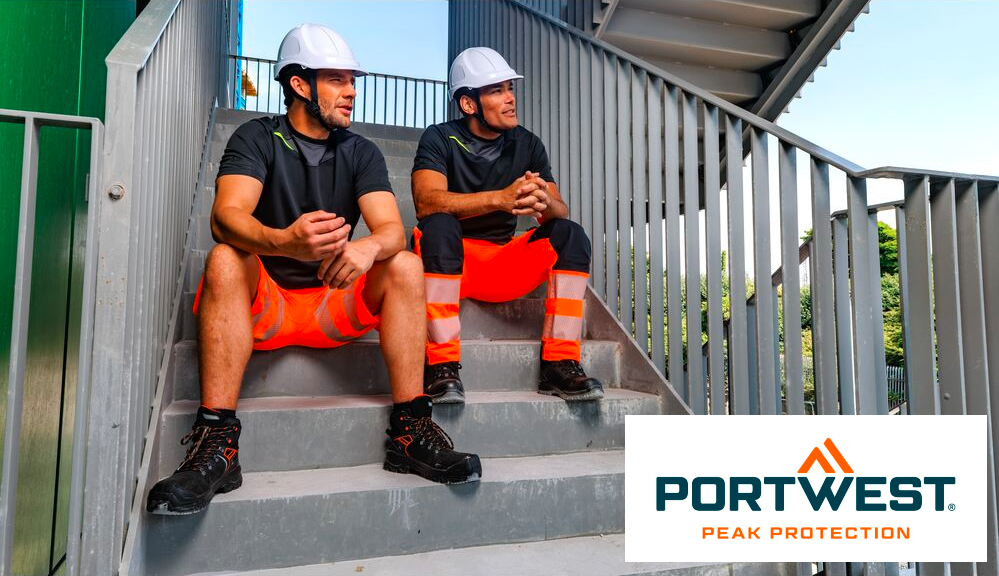 Due lavoratori con pantaloni arancioni ad alta visibilità e magliette nere. Entrambi gli uomini indossano caschi di sicurezza bianchi e sono seduti su una scala con una ringhiera grigia. Sullo sfondo si vedono il cielo azzurro e gli alberi, nell'angolo in basso a destra dell'immagine si può vedere il logo del produttore Portwest in lettere blu e arancioni su sfondo bianco. Ti forniamo un collegamento ai nostri pantaloni da lavoro.