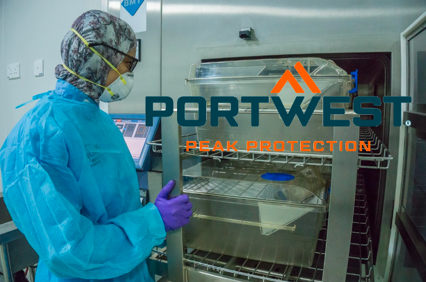 Persona con abbigliamento monouso blu da laboratorio davanti a un armadio in acciaio inox. Il logo del produttore Portwest in arancione e blu si trova a destra. Un link alla nostra sezione abbigliamento da laboratorio è incluso.