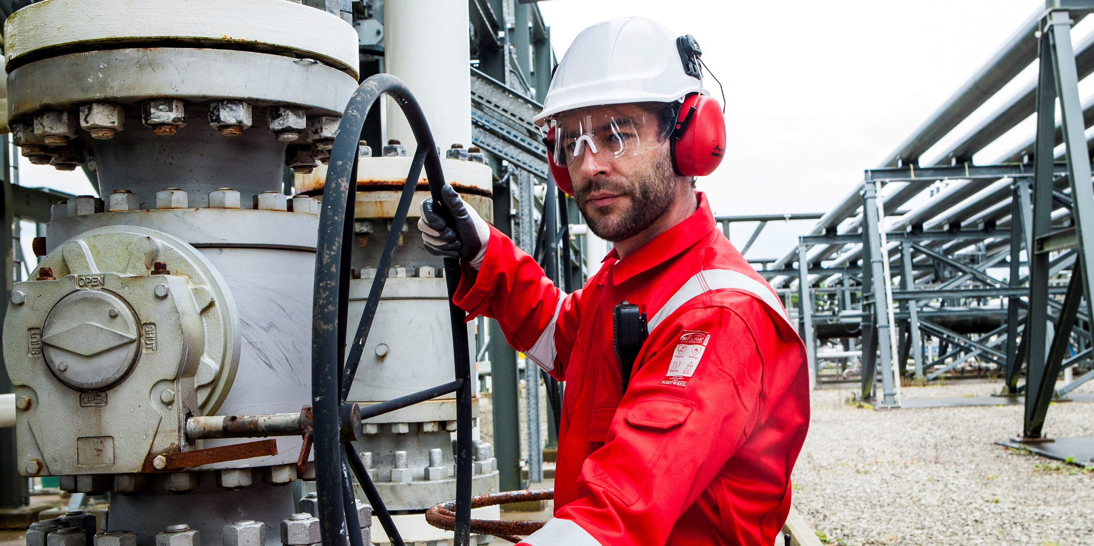 Operaio in un contesto industriale che indossa indumenti protettivi: casco bianco, occhiali, cuffie rosse, guanti neri e tuta FR50 rossa con strisce riflettenti.