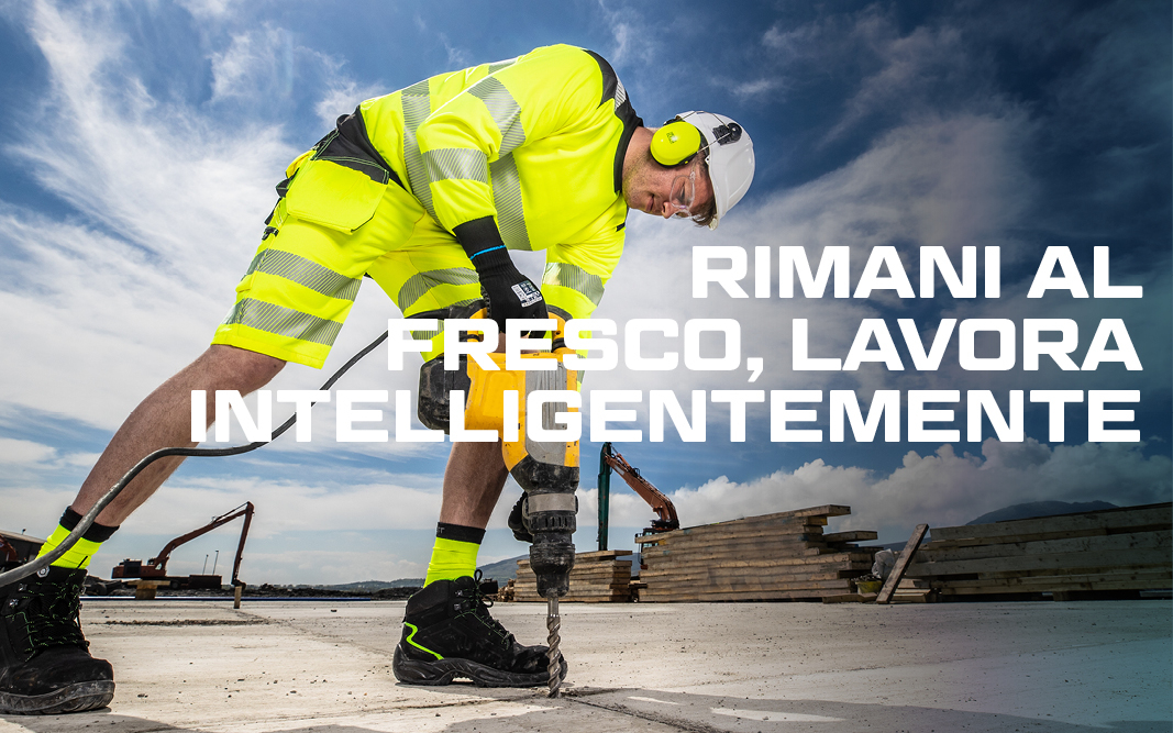 Lavoratore con casco e protezione dell'udito in abiti da lavoro estivi gialli ad alta visibilità che lavora con un martello pneumatico, sullo sfondo blu, cielo leggermente nuvoloso, escavatore e pila di assi di legno.