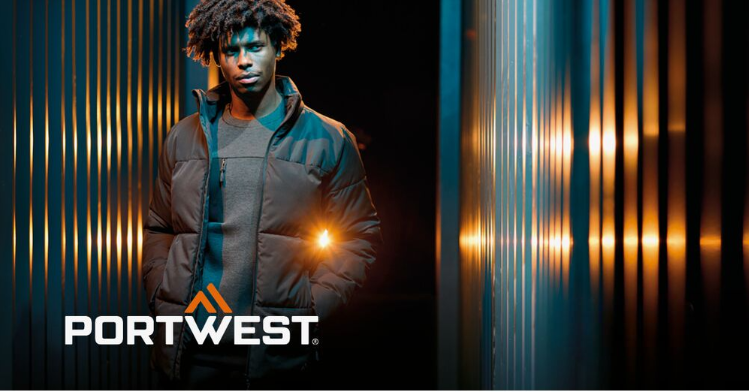 Un lavoratore in abiti da lavoro neri su uno sfondo astratto con una parete a strisce e riflessi di luce. Il logo del produttore, Portwest, in bianco e arancione, si trova nell'angolo in basso a sinistra dell'immagine.