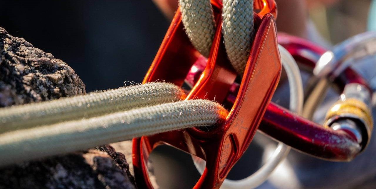 Primo piano di un sistema di corde arancioni con moschettoni rossi e argento fissato a una parete rocciosa.