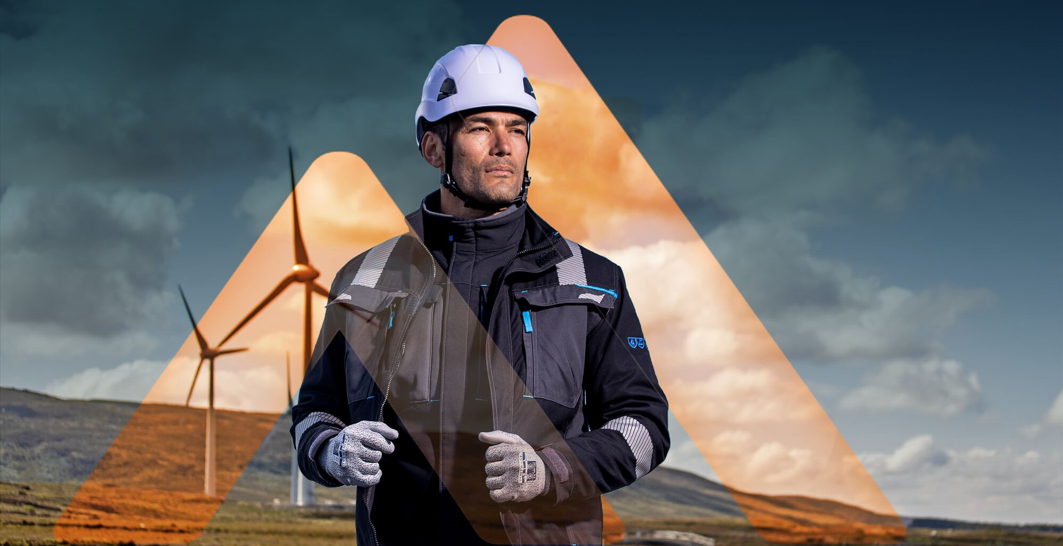 Un uomo con elmetto, guanti e giacca da lavoro si trova di fronte a turbine eoliche; illustrazione della sicurezza sul lavoro e della protezione anticaduta durante il lavoro in quota.