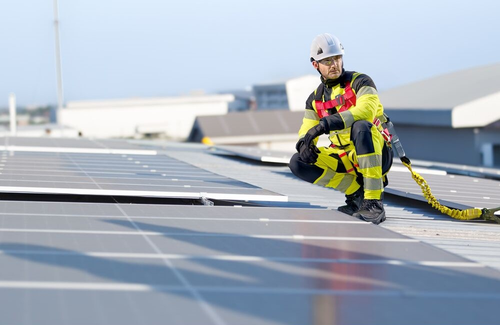 Lavoratore accovacciato su un tetto tra impianti fotovoltaici. Indossa abbigliamento da lavoro giallo ad alta visibilit&agrave;, un casco bianco, guanti e scarpe nere. &Egrave; assicurato con l&rsquo;imbracatura Portwest 3 punti Plus FP18. &Egrave; presente un link alla nostra protezione anticaduta.