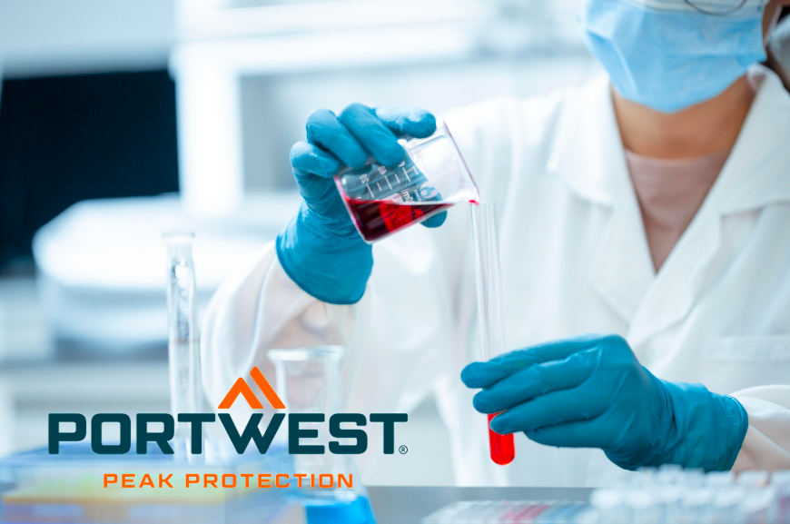Un addetto al laboratorio, in abiti protettivi con camice, guanti blu e mascherina, versa un liquido rosso da un becher in una provetta. Logo Portwest con lo slogan "Peak Protection" in primo piano. Viene fornito un collegamento alla nostra selezione di camici da laboratorio.