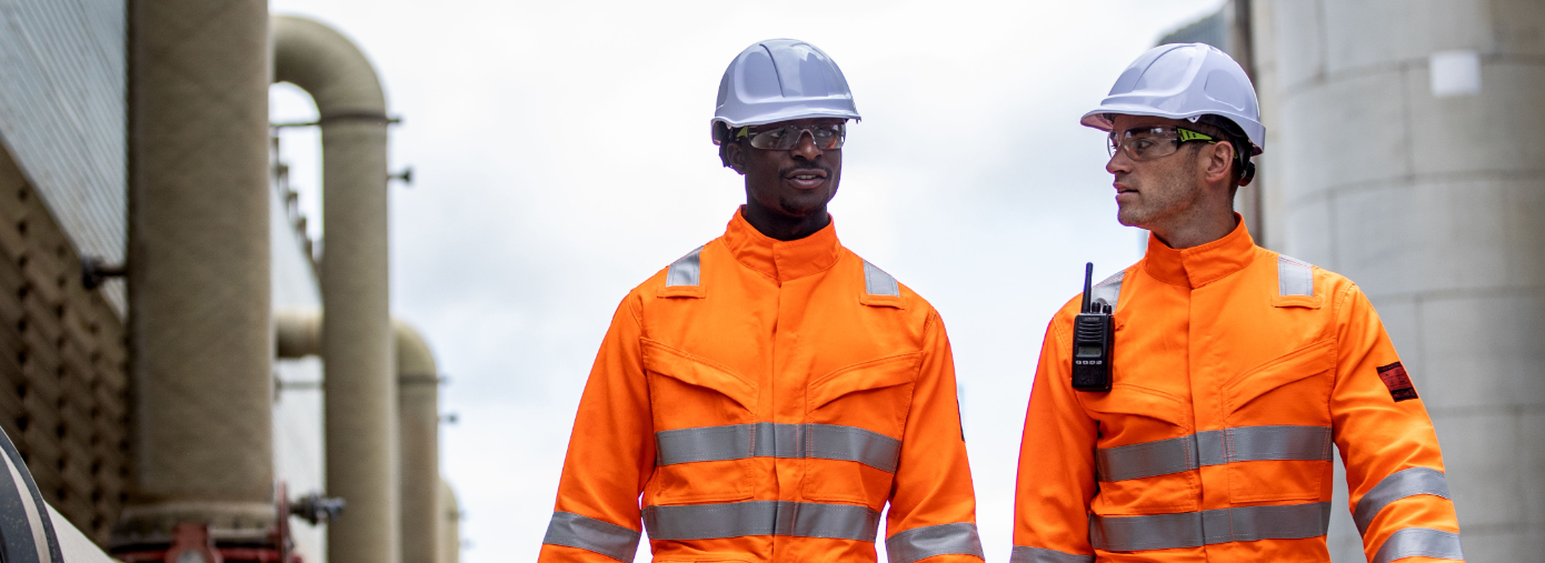 Due lavoratori di fronte a uno sfondo industriale che indossano indumenti arancioni ad alta visibilità ed elmetti bianchi.