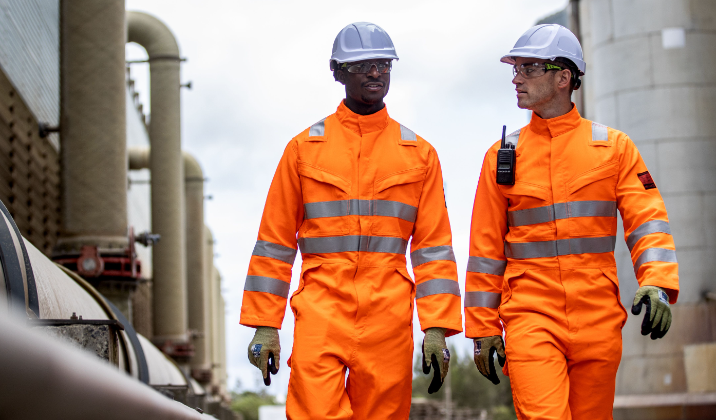 Due lavoratori di fronte a uno sfondo industriale che indossano indumenti arancioni ad alta visibilità ed elmetti bianchi. È disponibile un collegamento al casco Endurance Glowtex PG54.