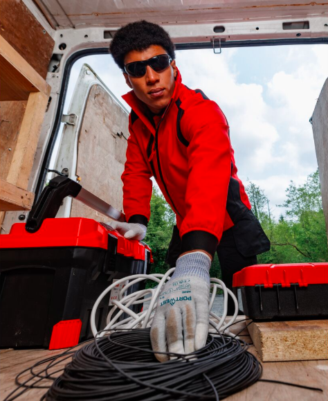 Un operaio con occhiali da sole, giacca softshell rossa, pantaloni cargo neri e guanti bianchi si infila all'interno di un furgone bianco per afferrare un fascio di cavi neri arrotolati. Sullo sfondo, si vede un cielo azzurro con nuvole e alberi verdeggianti. Un link conduce alla nostra selezione di guanti ESD.