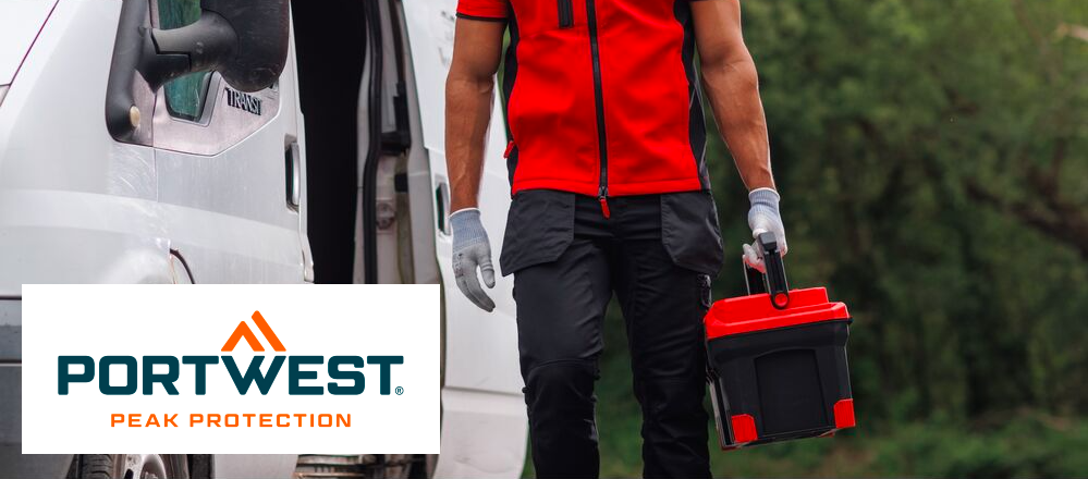Si può vedere la zona tra le spalle e i polpacci di una persona in abiti da lavoro. L'uomo indossa un gilet softshell rosso, una maglietta rossa sotto, pantaloni neri con tasche e guanti bianchi. Tiene una cassetta degli attrezzi nella mano sinistra. Alla sua sinistra, sullo sfondo, si vede un furgone bianco, il resto dello sfondo è coperto da alberi decidui verdi. Il logo blu e arancione del produttore Portwest su sfondo bianco si trova nell'angolo in basso a sinistra dell'immagine.