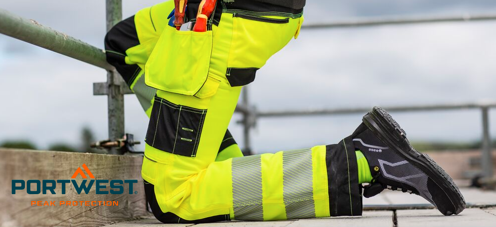 Un operaio edile è inginocchiato sul pavimento di un'impalcatura. Indossa pantaloni da lavoro ad alta visibilità giallo brillante con ginocchiere integrate e scarpe antinfortunistiche nere. Sullo sfondo si vede un cielo nuvoloso. Il logo del produttore, Portwest, in lettere blu e arancioni, è nell'angolo in basso a sinistra.