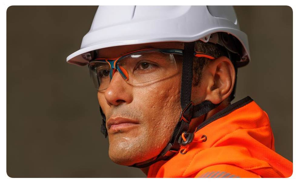 Ritratto di un uomo con abbigliamento ad alta visibilità arancione, casco di protezione bianco e occhiali di sicurezza. È presente un link alla nostra collezione di occhiali.