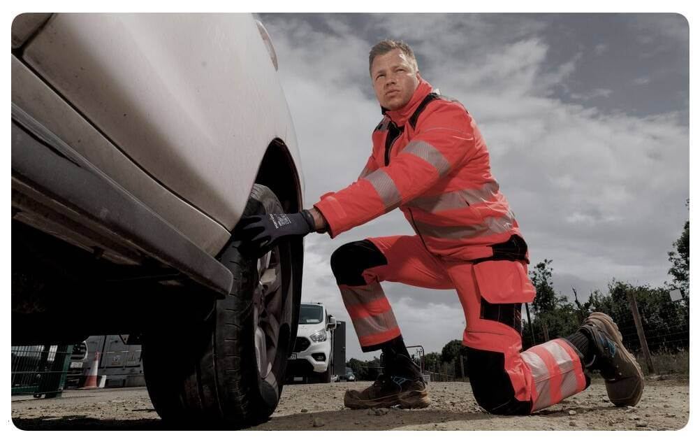 Un uomo con abbigliamento ad alta visibilit&agrave; arancione &egrave; inginocchiato davanti a un'auto bianca e lavora su una ruota. Sullo sfondo si vede un cielo nuvoloso. &Egrave; presente un link alla nostra collezione di protezioni per le ginocchia.