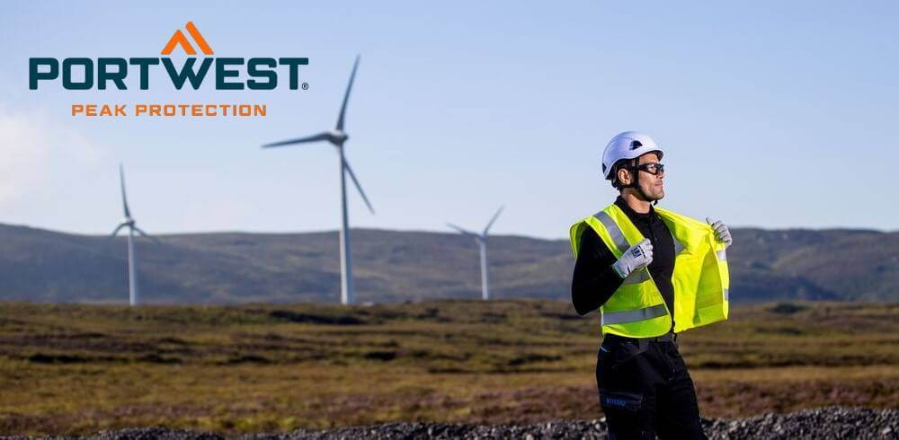 L'immagine mostra un uomo in abiti da lavoro neri in piedi davanti a un paesaggio brullo con tre turbine eoliche. Indossa un elmetto bianco, un giubbotto di sicurezza giallo, guanti e occhiali di sicurezza. Il logo Portwest è visibile in blu e arancione nell'angolo in alto a sinistra dell'immagine.