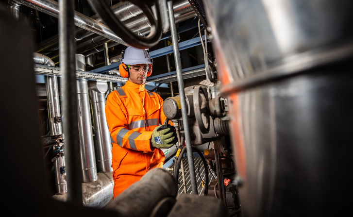 Lavoratore con casco bianco e protezione per le orecchie in abiti da lavoro arancioni con guanti su sfondo industriale. L'immagine è legata alla collezione di guanti da lavoro.