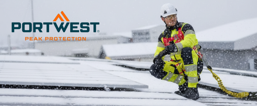 Un operaio con una tuta gialla ad alta visibilit&agrave; con strisce riflettenti &egrave; inginocchiato su un tetto innevato accanto a un impianto fotovoltaico. Indossa un casco bianco e una protezione anticaduta. Sullo sfondo si vedono altri tetti innevati e il logo del produttore, Portwest, &egrave; visibile in blu e arancione in alto a sinistra dell'immagine.