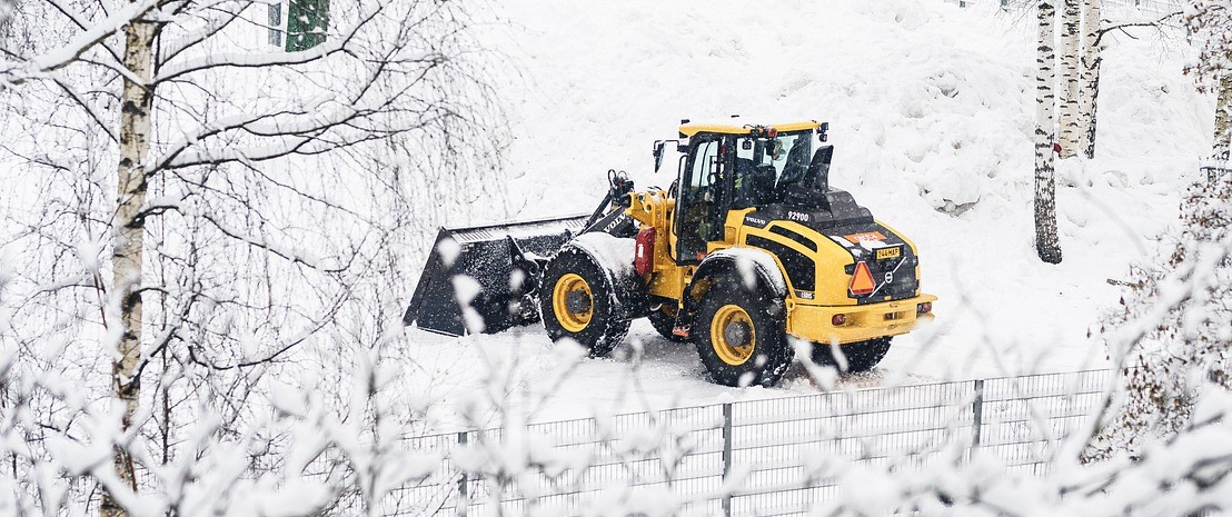 Trattore giallo con pala in paesaggio innevato con recinzioni e betulle.